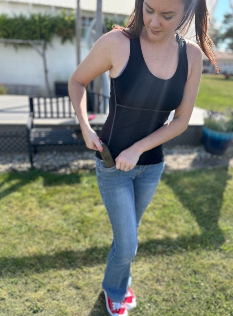Woman in black tank top and blue jeans standing outdoors on grass with king sized travel case in pocket