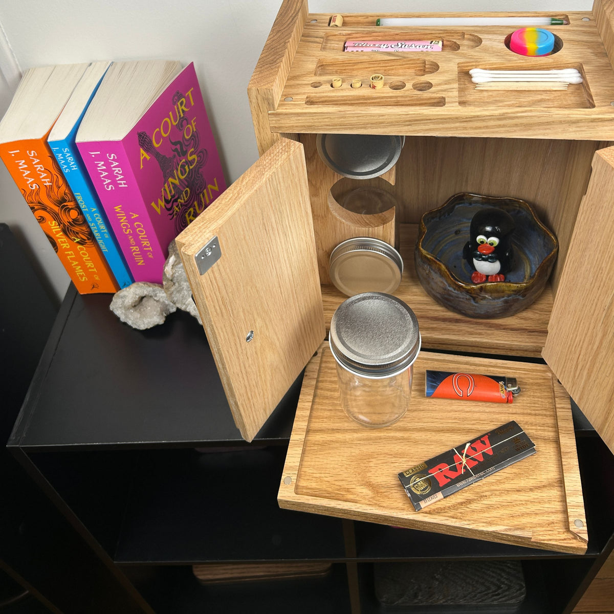 A light natural wood storage cabinet with multiple open compartments, shown holding glass jars with metal lids, a bowl, a lighter, and a RAW rolling paper pack. The cabinet is placed on a black shelf next to books.